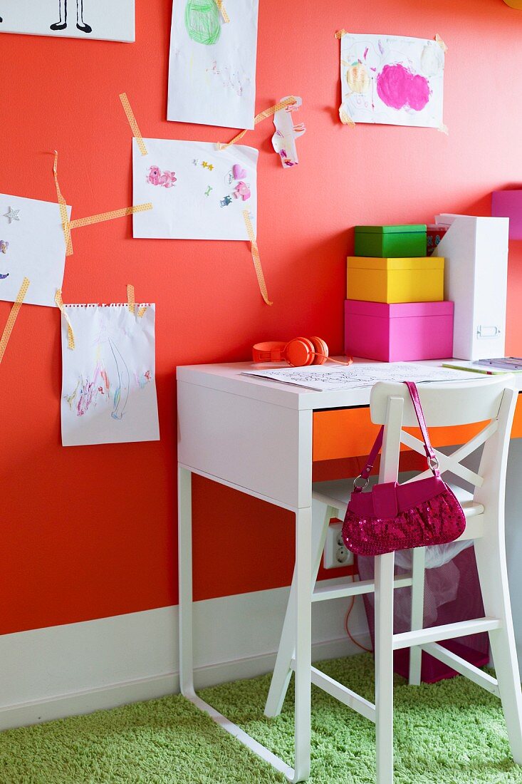 Weisser Hochstuhl vor Schreibtisch an orangeroter Wand mit Kinderzeichnungen
