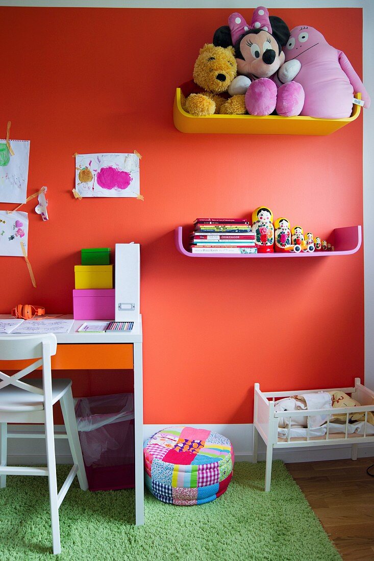 Farbige Ablagen mit Kuscheltieren und Büchern an orangeroter Wand, seitlich Schreibtisch im Kinderzimmer