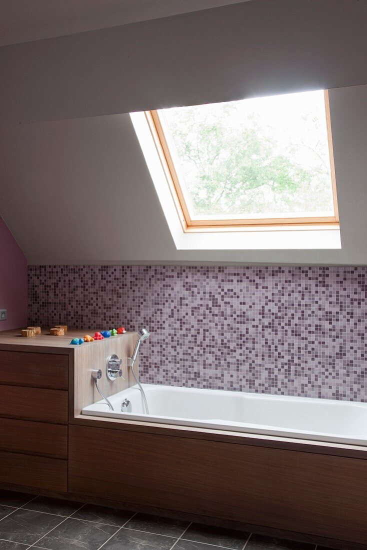 Bathtub with wooden fittings and mosaic-tiled side wall under window in sloping ceiling