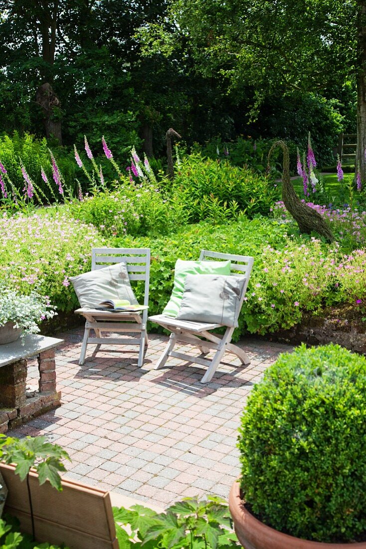 Holzklappstühle mit Kissen auf einer Terrasse, dahinter Garten mit violetten Lupinenblüten und Vogelskulpturen