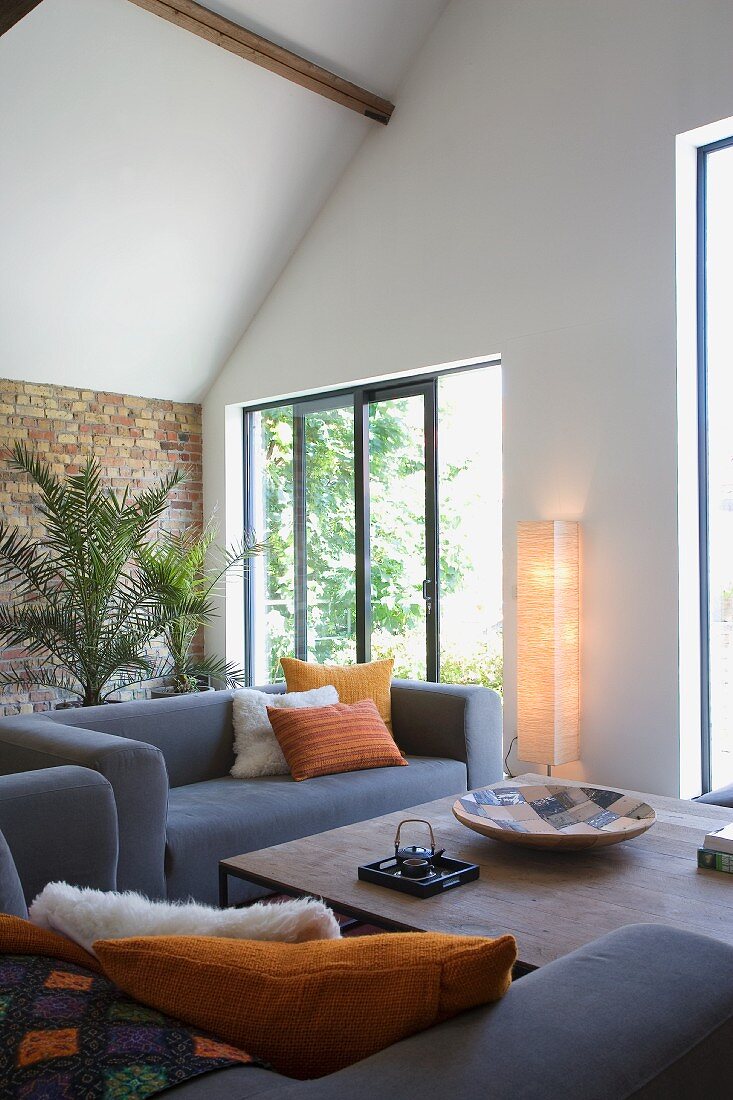 Loungebereich mit grauem Sofa und Couchtisch, im Hintergrund Terrassentür zwischen Zimmerpalme und Stehleuchte mit Papierschirm