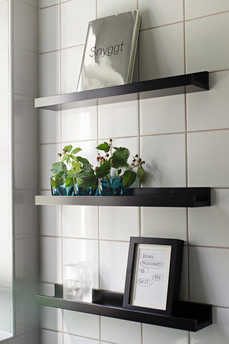 Vases of leaves and framed picture on black floating shelves on white-tiled wall