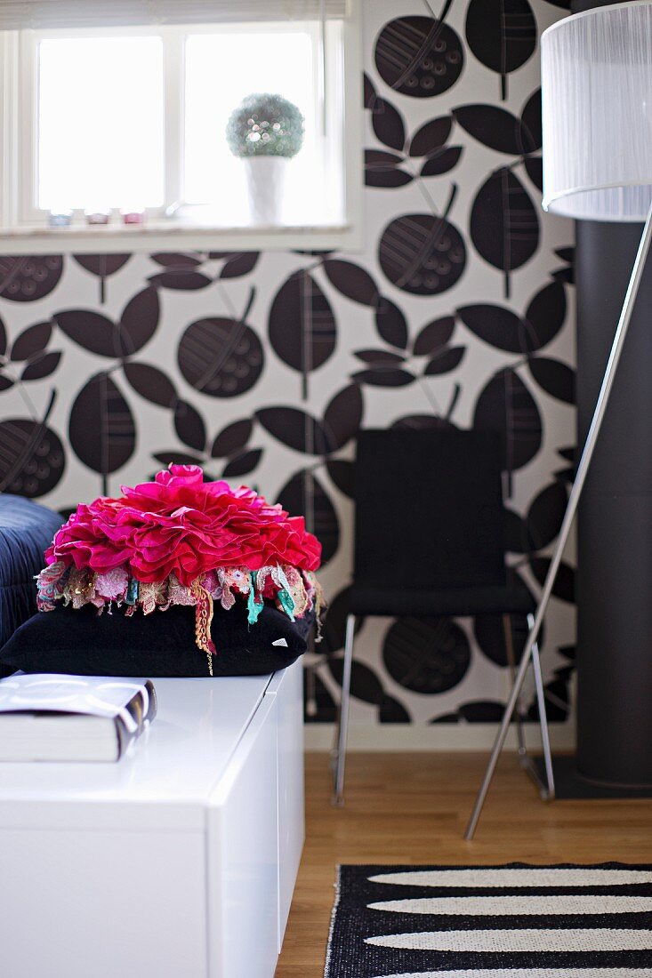 Pinkfarbenes Kissen in Blütenform auf weißem Sideboard, im Hintergrund schwarz-weisses Blumenmuster auf Tapete an Wand