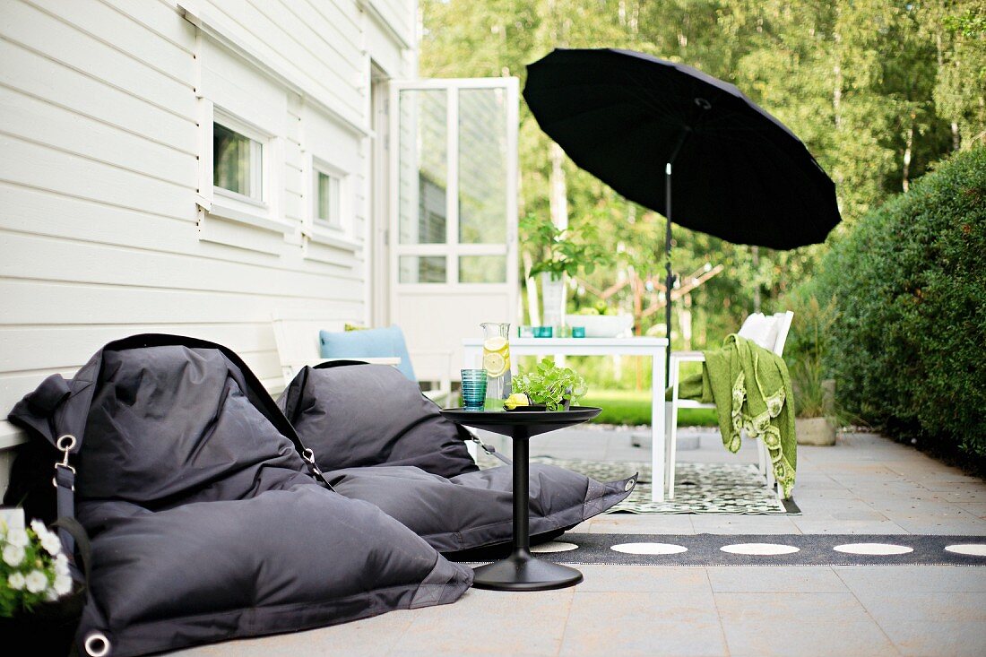 Schwarze Sitzsäcke um Beistelltisch, im Hintergrund Tisch unter schwarzem Schirm auf Terrasse, sonnenbeschienener Garten
