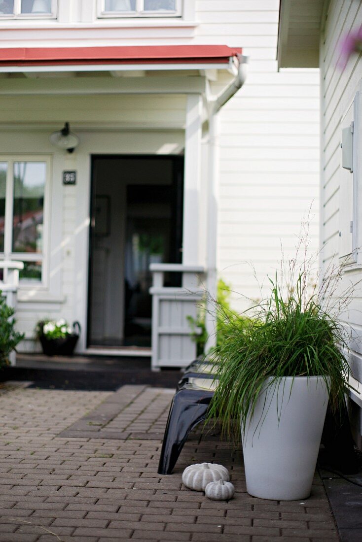Ornamental grass in white planter on terrace in front of white clapboard house with porch and open door