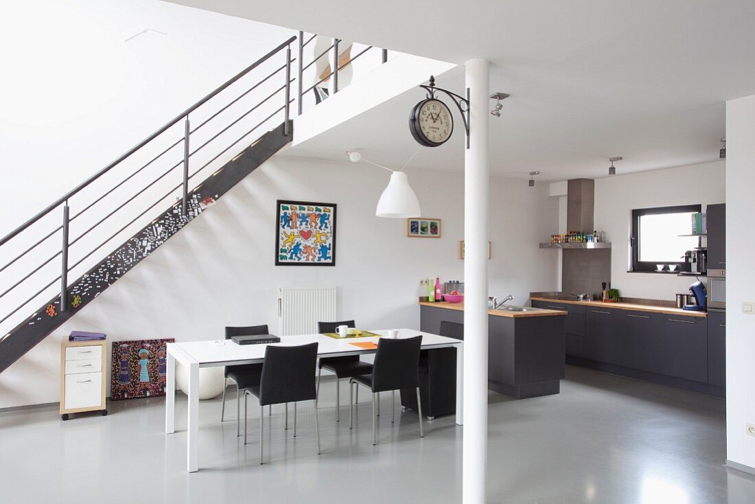 Minimalistische Loftwohnung mit Retro Charme, weisser Tisch und schwarze Stühle vor Treppe aus Metall, im Hintergrund offene Küche mit dunkelgrauen Schrankfronten