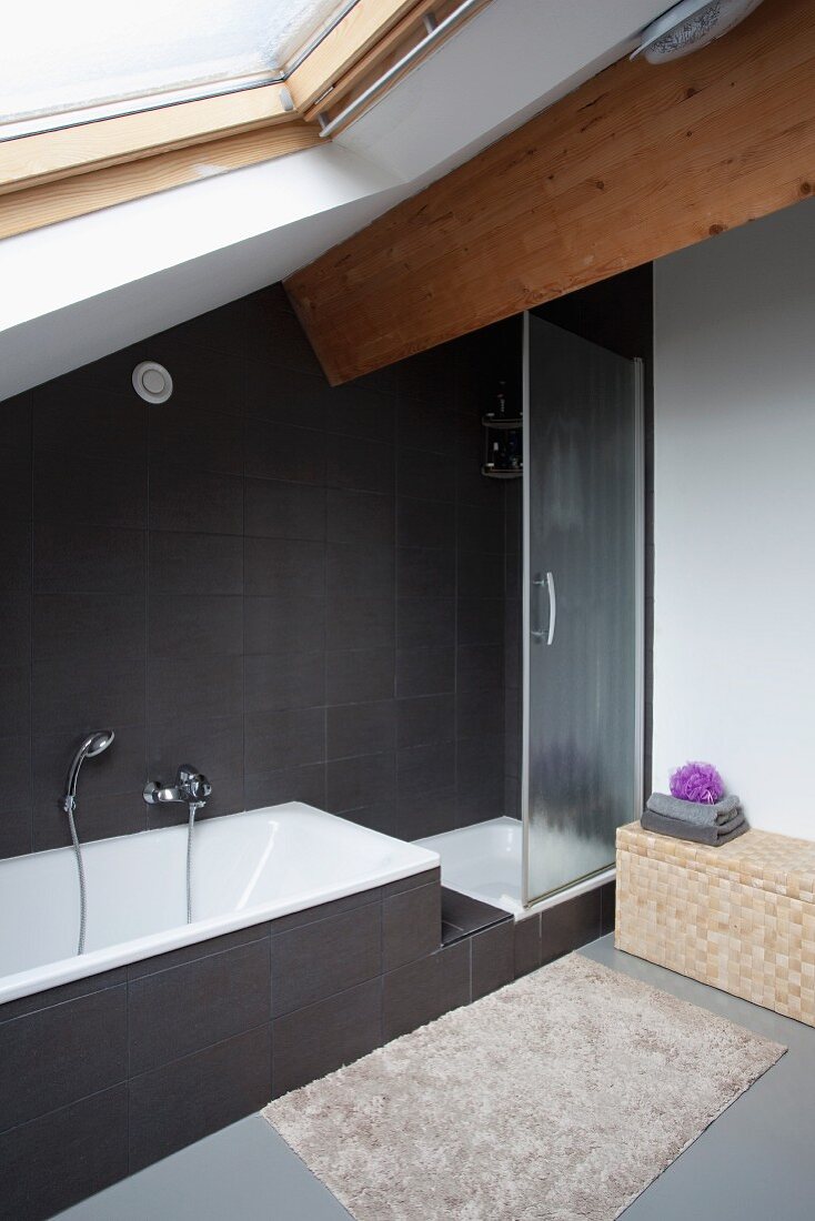 Designer, attic bathroom with fitted bathtub and shower cubicle against black-tiled wall