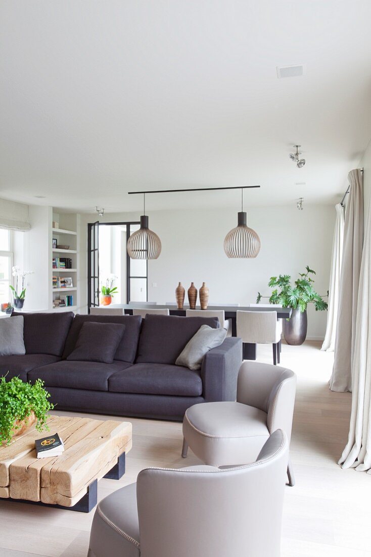 Graue Polstersessel im Fiftiesstil und Sofa um Bodentisch aus rustikalen Vierkanthölzern in minimalistischem Wohnraum, im Hintergrund skandinavische Designerleuchten über Essplatz