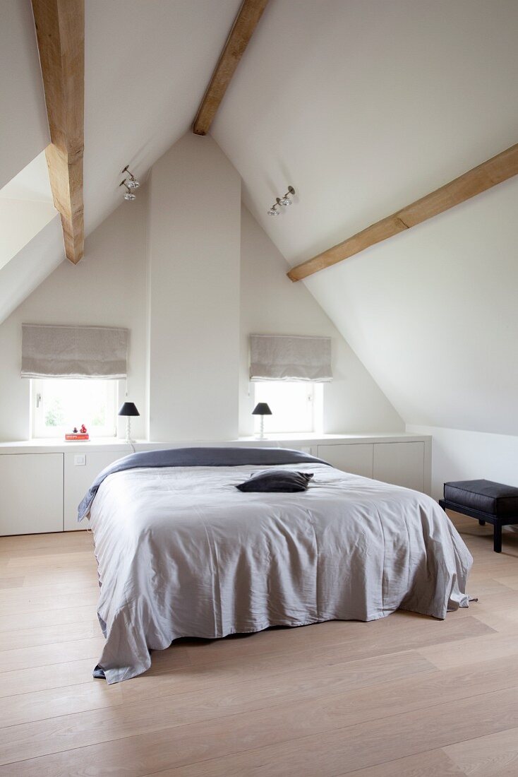 Sleeping area in converted, minimalist attic; simple double bed against gable-end wall with windows