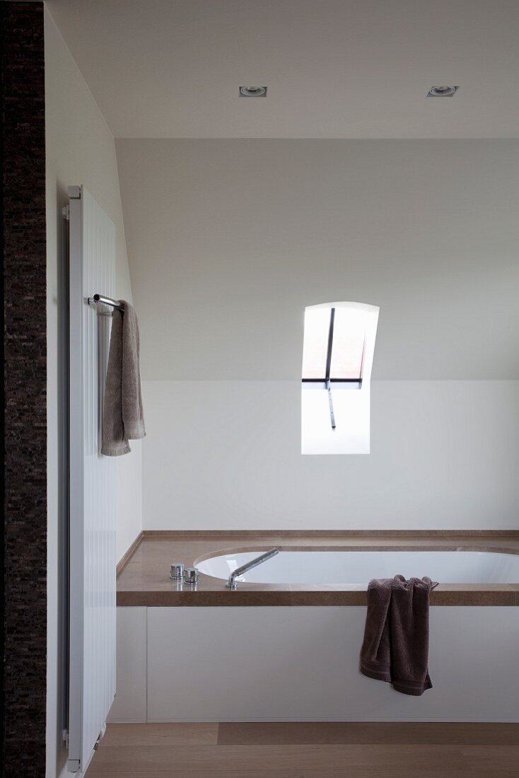 Fitted bathtub with dark stone surround under sloping ceiling with small pivot-hung window