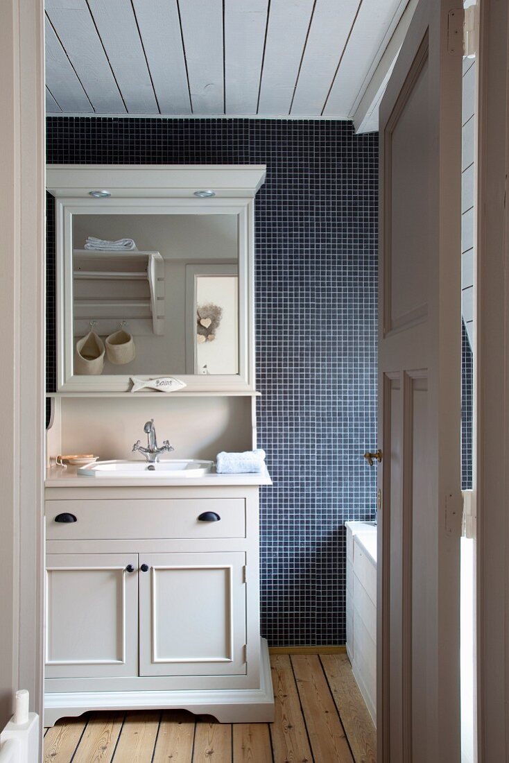 Blick durch offene Tür auf weissen Holzschrank mit Spiegelaufsatz und eingebautem Waschbecken vor dunkler Mosaikfliesenwand