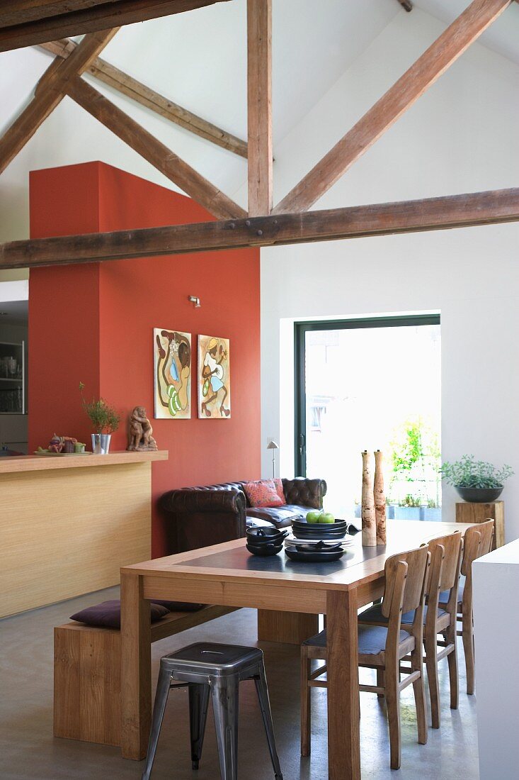 Retro stools and chairs at wooden table opposite counter adjoining red-brown wall in rustic, modern interior