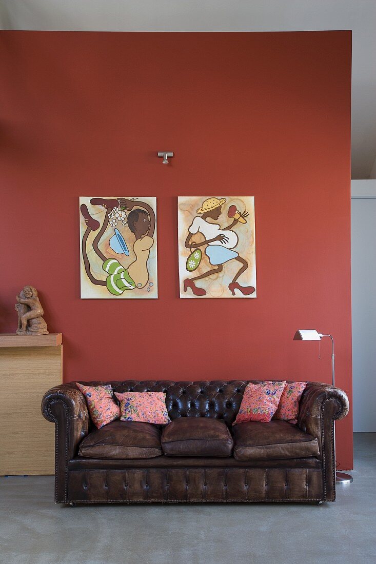 Brown, vintage leather couch below artworks on red-brown wall in modern interior