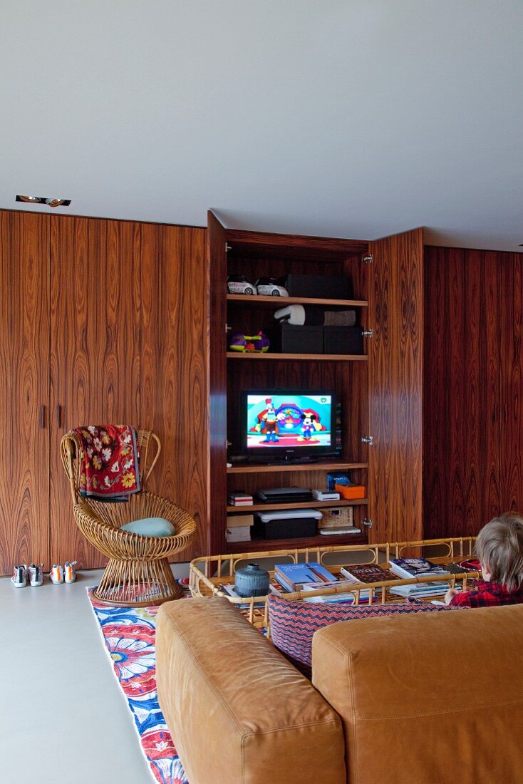 Ausschnitt eines Loungebereiches, Rattan Sessel vor Einbauschrank aus Palisanderholz mit Fernseher hinter offener Tür