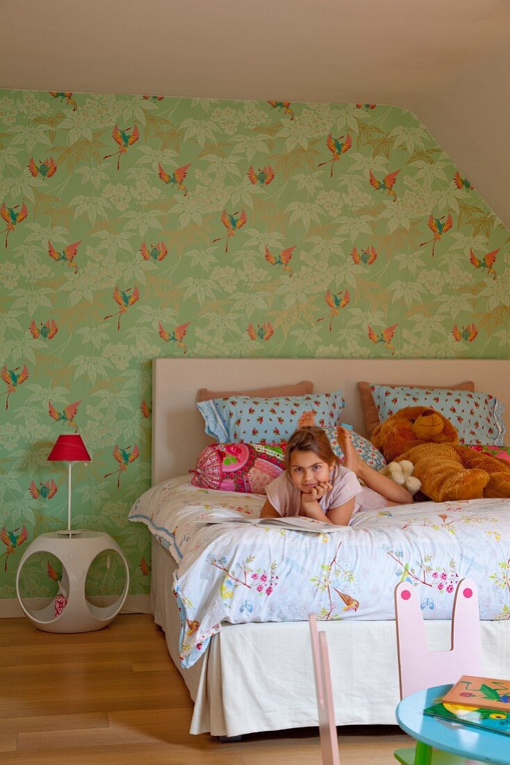 Mädchen beim Lesen auf dem Bett, daneben Retro Nachttisch mit Tischleuchte vor grün tapezierter Wand mit floralem Motiv