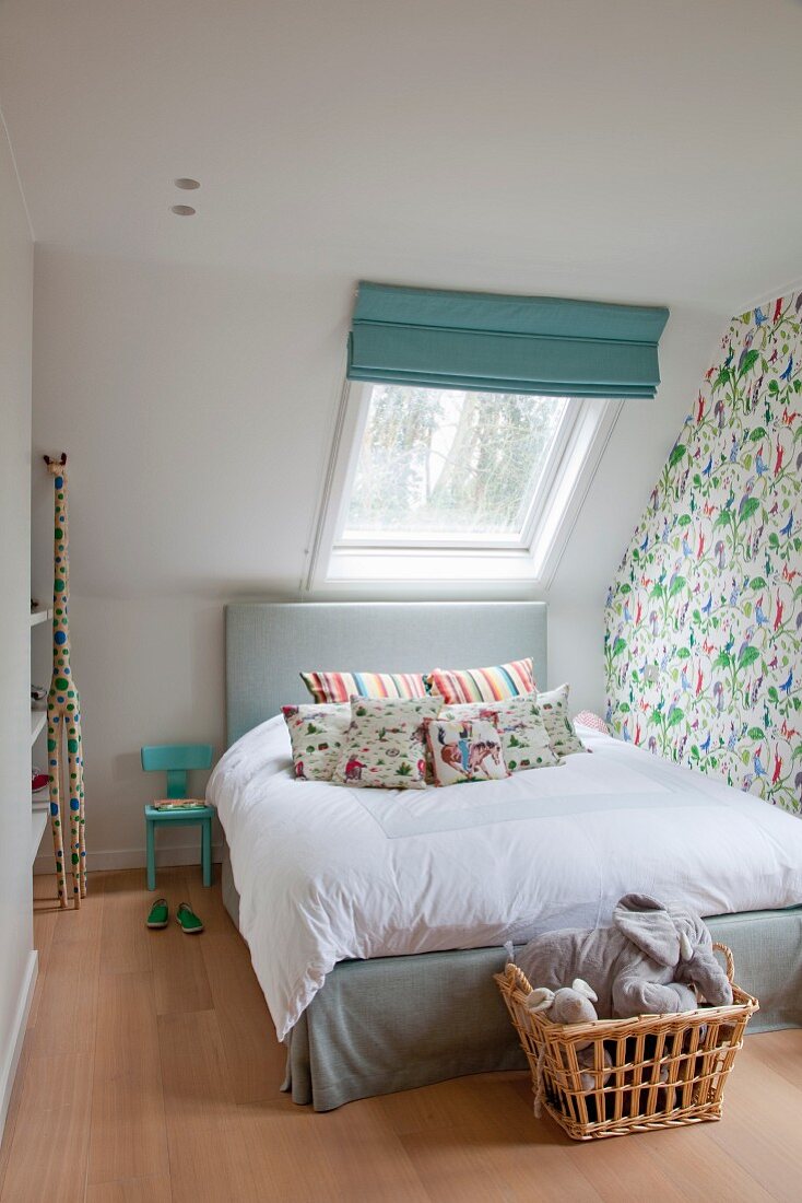 Bett mit gepolstertem Kopfteil vor Dachflächenfenster in Kinderzimmer, Wäschekorb mit Stofftieren an Bettende