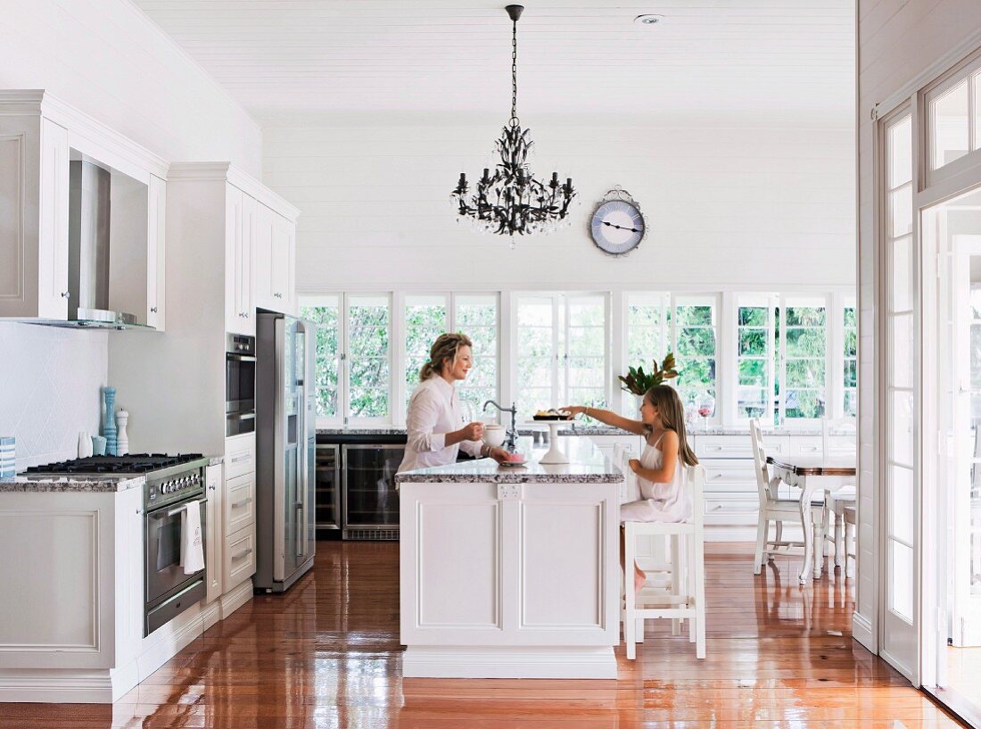 Woman and girl seating at country-house-style island counter below Rococo chandelier in open-plan kitchen