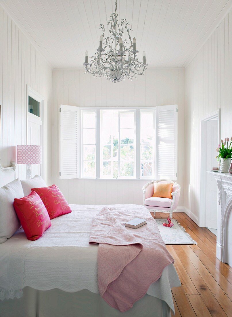 Doppelbett mit roten Kissen auf weisser Bettwäsche, darüber Rokoko-Kronleuchter in weißem, holzverkleidetem Schlafzimmer