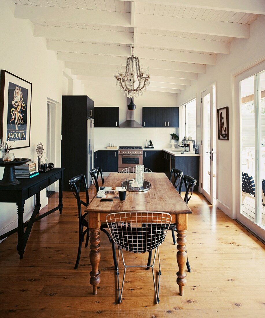 Verschiedene Klassikerstühle um altem Holz Esstisch in offenem Wohnraum, im Hintergrund moderne Küche in Schwarz
