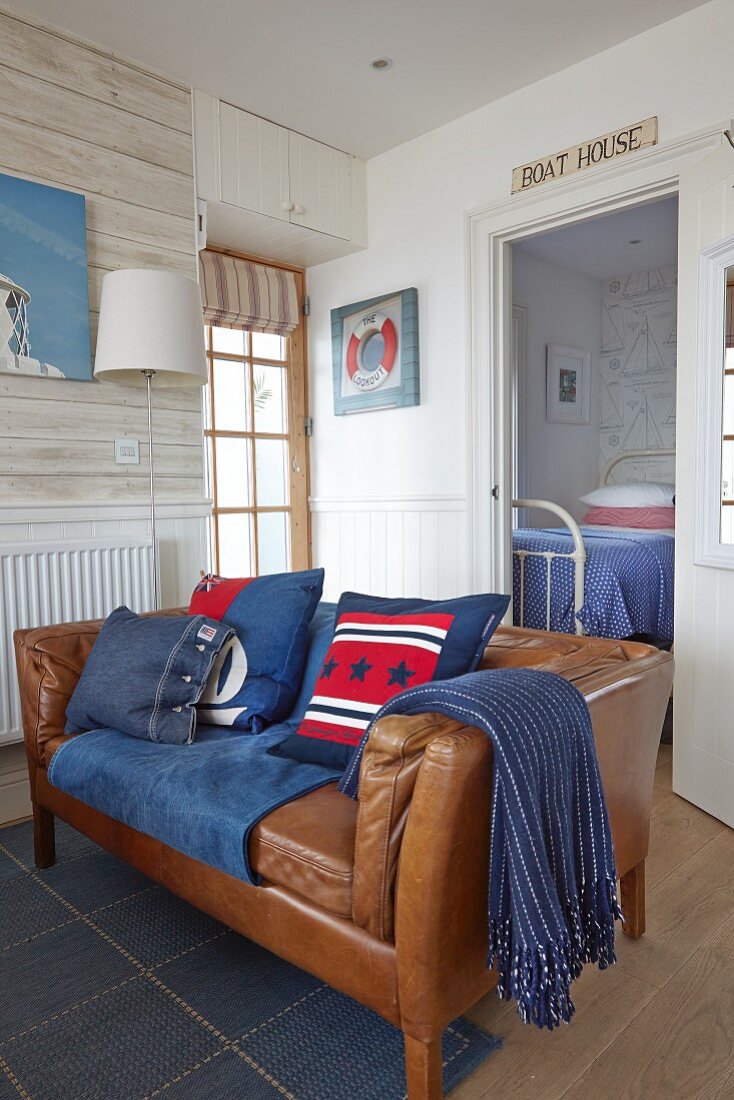 Braunes Ledersofa mit Kissen in blauen und roten Tönen und nautische Accessoires im Wohnzimmer mit Tapete in Holzoptik