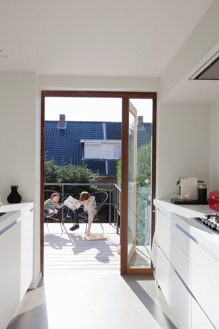 Sonnenbeschienene weiße Designerküche mit offener Balkontür und Kindern auf Stühlen im Freien