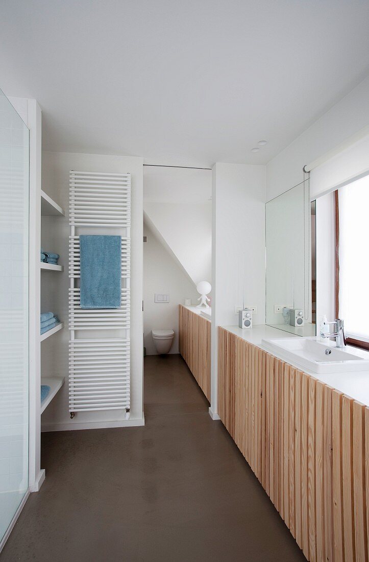 Modern bathroom; fitted cupboards with vertical slatted doors and integrated sink below window, white towel warmer on wall next to sliding door