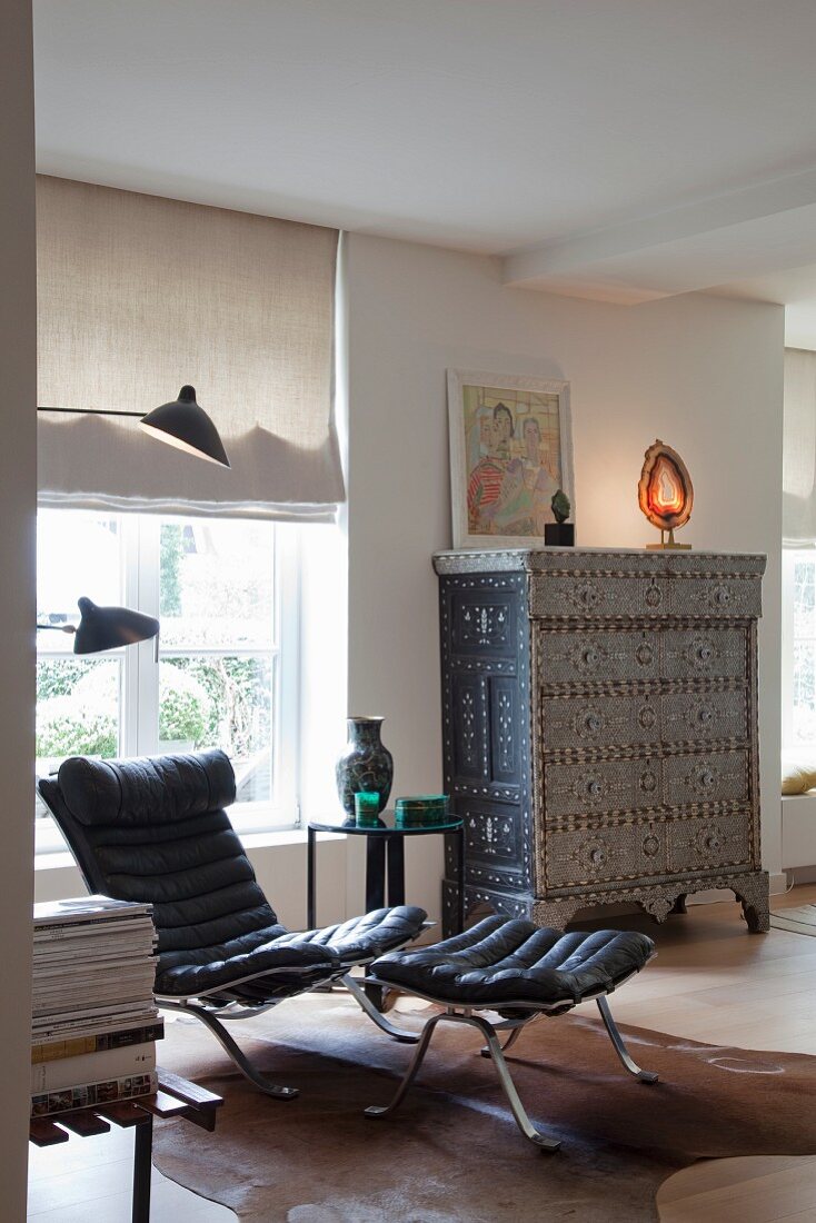 Black retro leather armchair with matching footstool in front of window; Oriental cabinet against wall in background