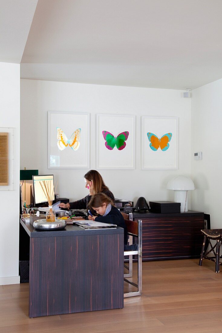 Frau mit Kind am Schreibtisch in Zimmernische, an Wand Bildergalerie mit buntem Schmetterlingmotiv