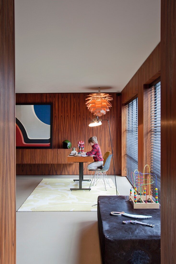 Blick durch Türöffnung auf Schreibtisch und Kind in elegantem Arbeitszimmer mit Palisander vertäfelten Wänden und Klassiker Hängeleuchte