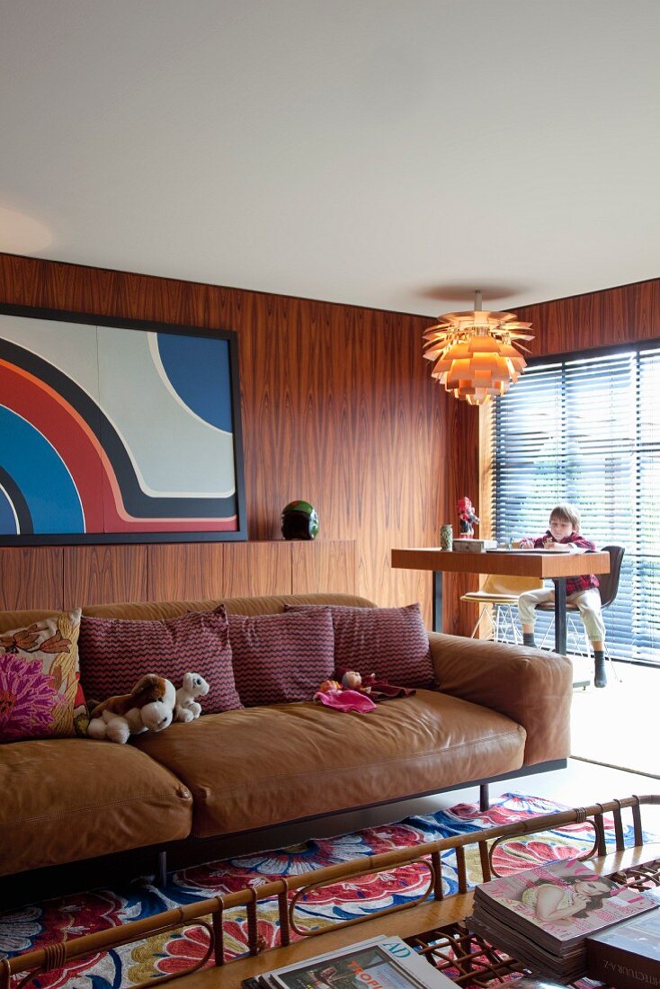 Brown leather couch with row of scatter cushions; child sitting at desk in background in corner of wood-panelled room