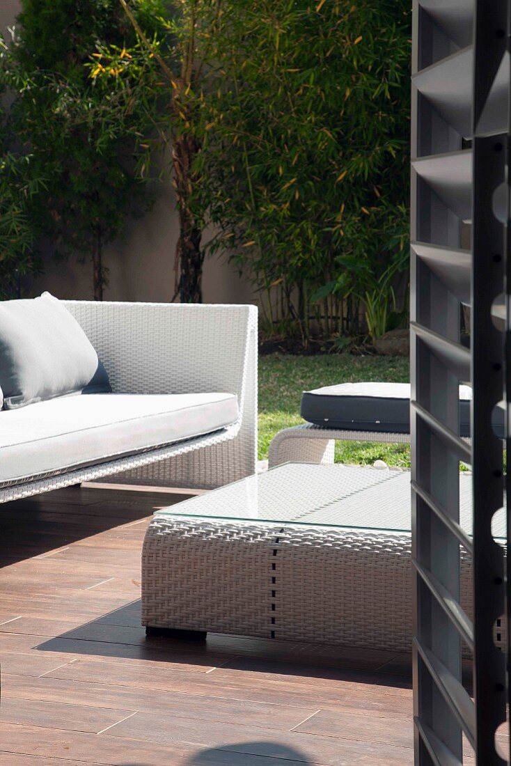 Blick durch offene Tür auf moderne Outdoormöbel aus weißem Rattan auf Holzterrasse und sommerlicher Stimmung