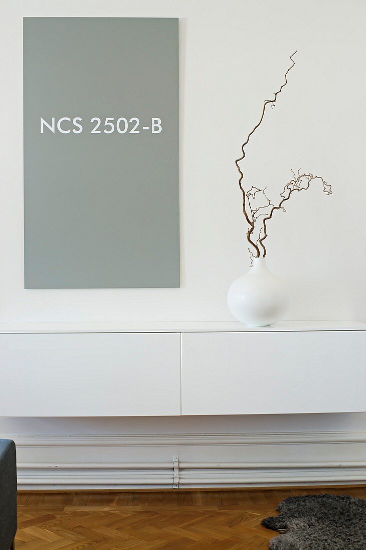 Vase mit Zweig auf minimalistischem Hängebord in Weiß, vor grauer Tafel an Wand mit Farbnummer des NCS Farbsystems