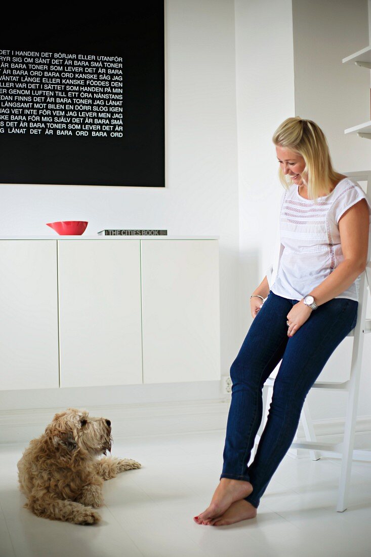 Frau mit Hund in minimalistischem Wohnraum, an Wand weisses Sideboard montiert unter schwarzer Tafel mit Text