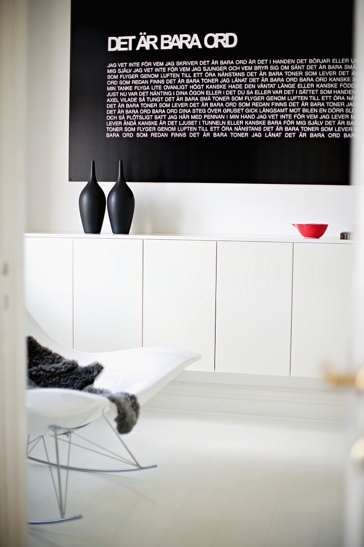 Teilweise sichtbarer moderner Schaukelstuhl und an Wand montiertes Sideboard in Weiß, darauf schwarze Gefässe vor schwarzer Tafel mit Text