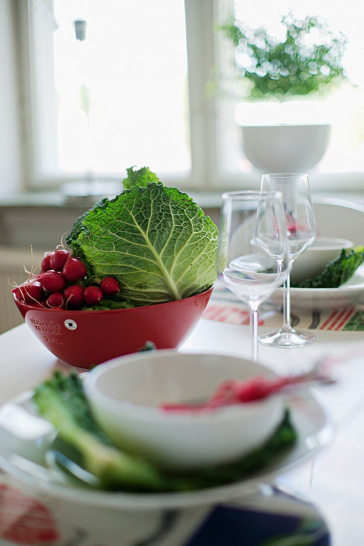 Im Vordergrund ein Gedeck mit weisser Schale, vor roter Schale mit Gemüse auf dem Tisch