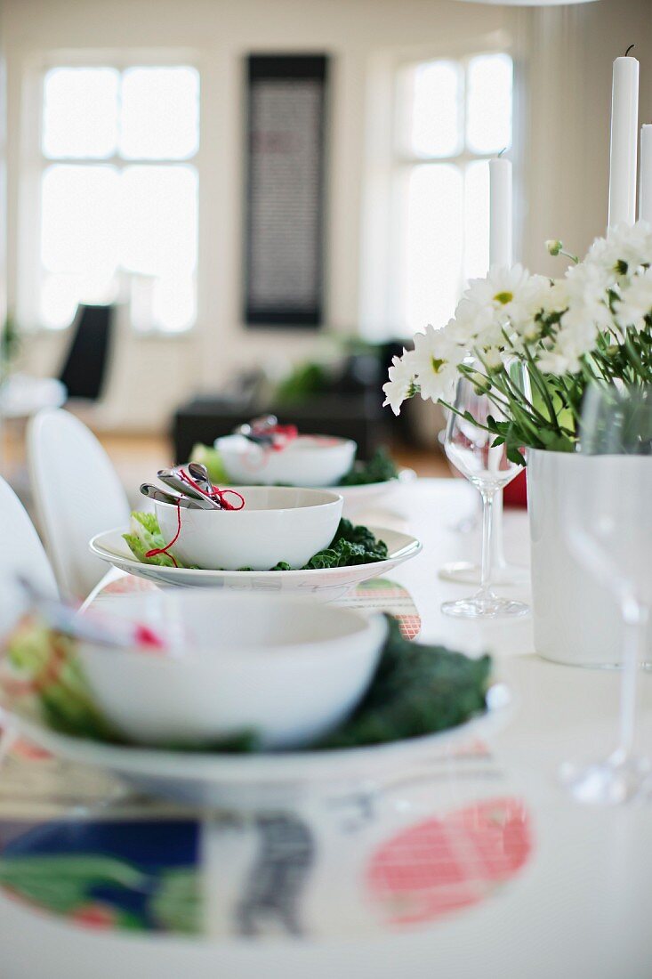 Gedecke mit weissen Schalen und Wirsingblatt dekoriert, teilweise sichtbarer Blumenstrauss in Vase auf Tisch