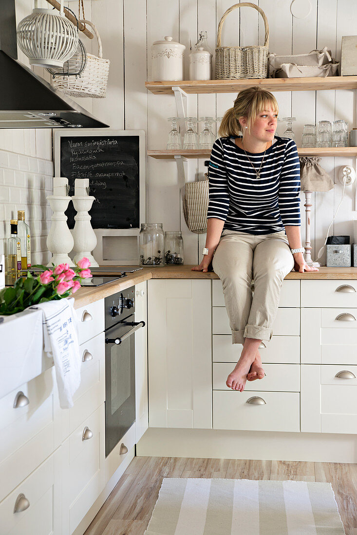Frau in weisser Landhausküche übereck, an Holzwand Konsolenregal mit Geschirr
