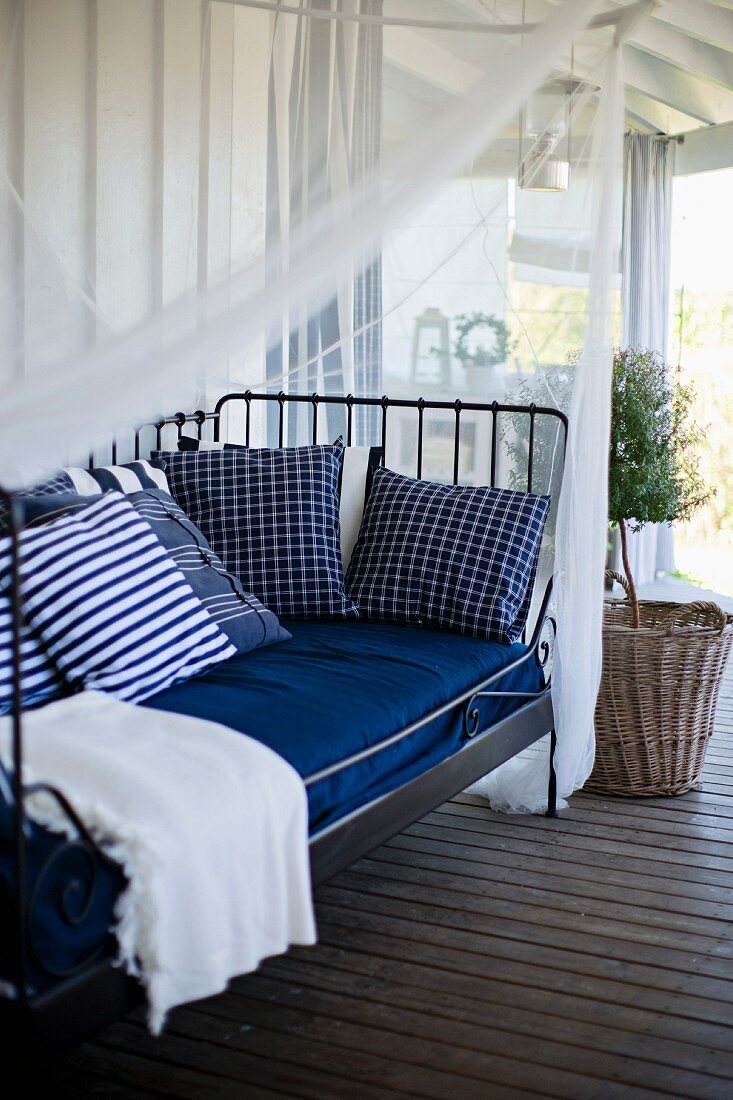 Metal day bed with white and blue cushions below canopy in front of small potted tree on wooden veranda floor