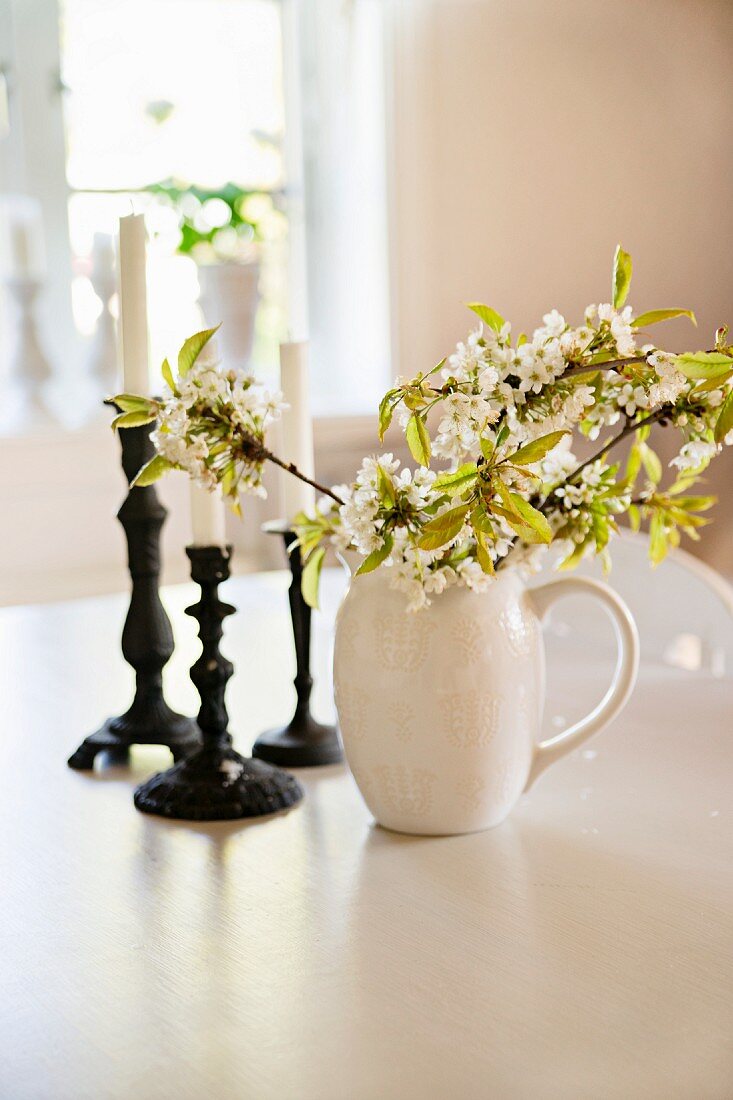 Zweige mit weissen Blüten in Porzellankrug und Kerzenständer auf weißem Tisch