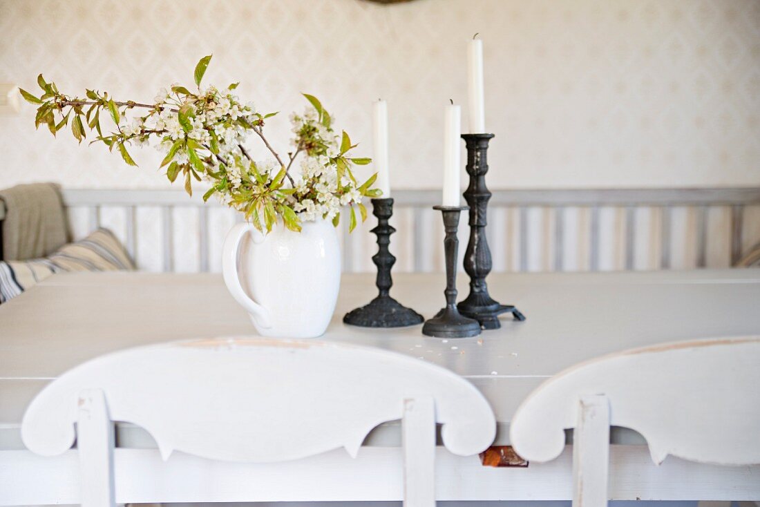 Porzellankrug mit Blätterzweig und Kerzenständer auf Tisch, vorne teilweise sichtbare, geschwungene Rückenlehne eines Küchenstuhls