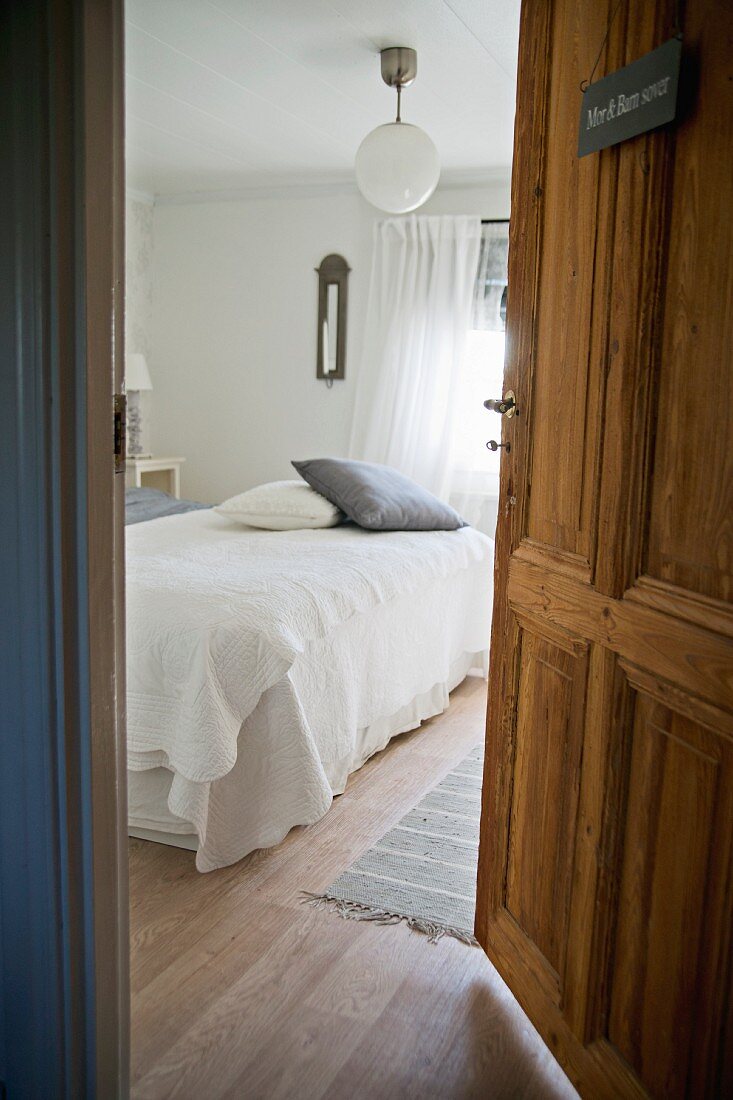 Blick durch offene Tür auf Bett mit weisser Tagesdecke und Kissen in ländlichem Schlafzimmer