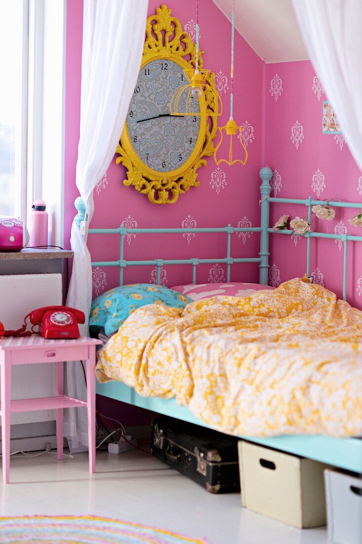 Kinderbett mit türkisfarbenem Retro Metallgestell an rosa tapezierter Wand, Uhr mit gelbem, verziertem Rahmen