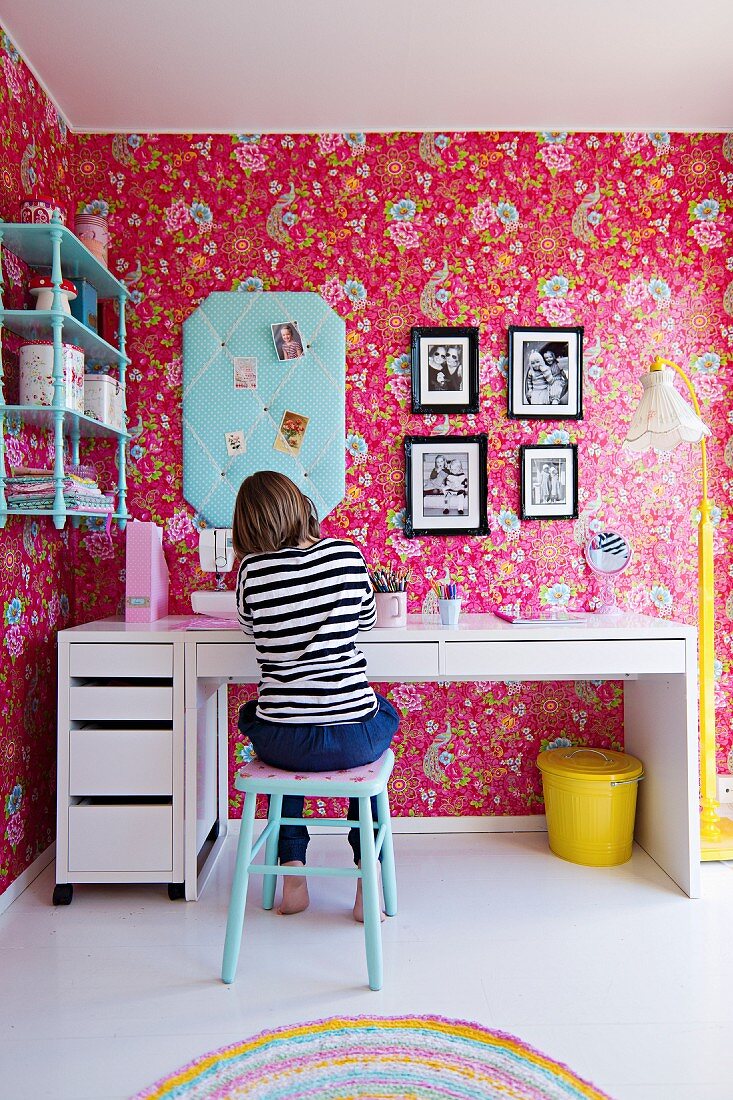 Junges Mädchen am Schreibtisch in nostalgischem Kinderzimmer, an Wand Tapete mit Blumenmuster auf pinkfarbenem Hintergrund