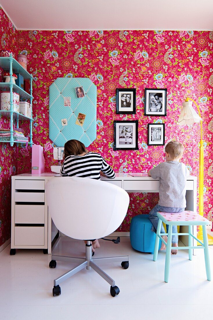 Mädchen und Junge am Schreibtisch in nostalgischem Kinderzimmer, an Wand Tapete mit Blumenmuster auf pinkfarbenem Hintergrund
