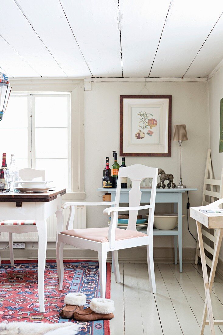 White-painted chairs next to table in front of window in rustic dining room with white board floor and ceiling