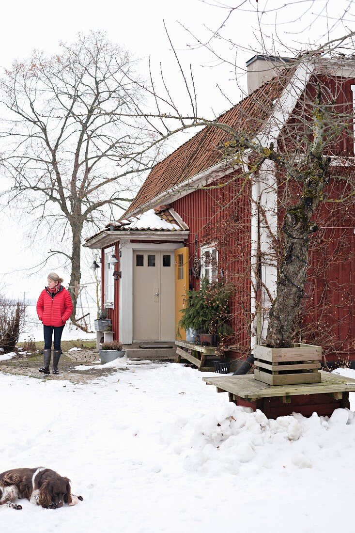 Frau und Hund in verschneitem Garten vor rotbraunem Holzhaus mit Windfang Eingang