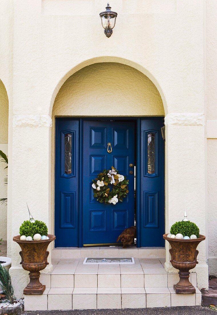 Blaue Haustür einer Villa mit Holzkassetten und aufgehängtem Weihnachtskranz, auf Treppenstufen bepflanzte Amphoren