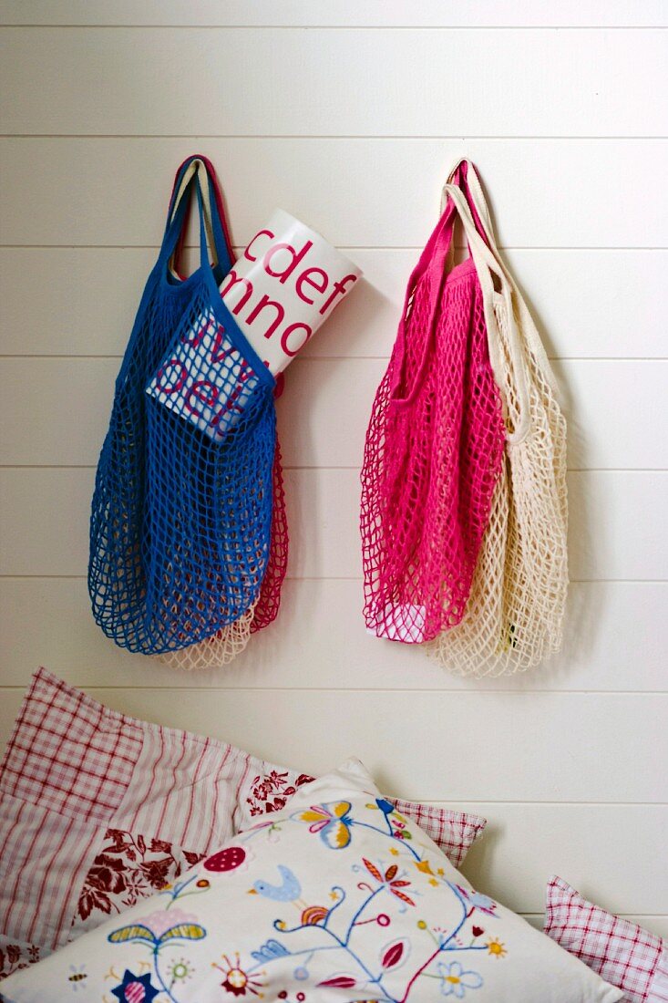 Veschiedenfarbige Netz Einkaufstaschen an weisser Holzwand aufgehängt, darunter besticktes Kissen