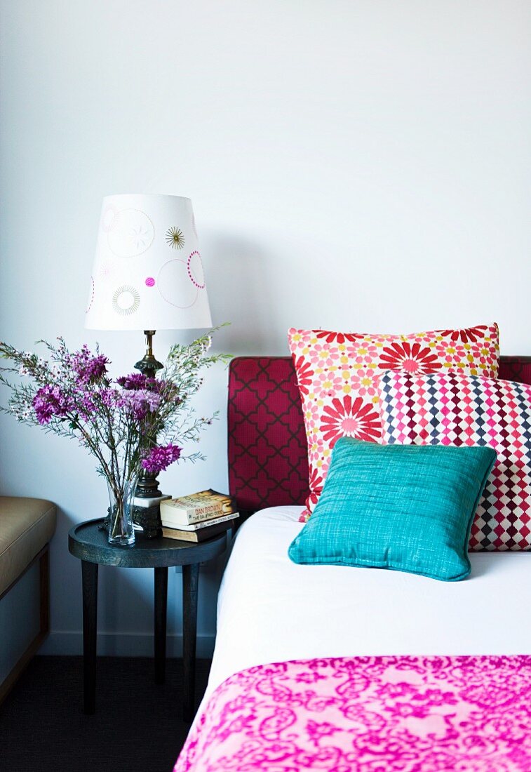 Stack of colourful scatter cushions on bed next to table lamp and vase of flowers on bedside table