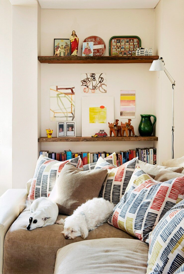 Two white dogs and many scatter cushions with graphic patterns on corner sofa in front of bookshelves in niche